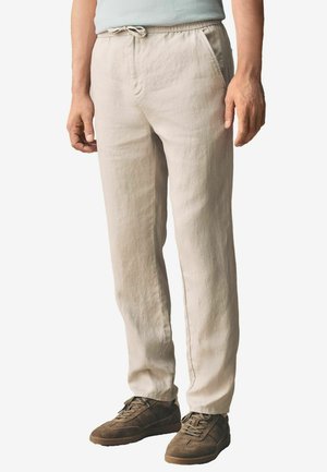 Beige linen pants with an elastic waistband and drawstring, featuring side pockets and a relaxed fit, paired with brown footwear.