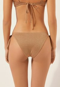 Bikiniunderdel i en metallisk beige färg, med en texturerad yta och justerbara sidoknytningar för en anpassningsbar passform.