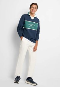 Pullover blu navale con una striscia verde; presenta un colletto, rifiniture bianche e il logo "Silbon Sports". Abbinato a pantaloni bianchi e sneakers blu navy.