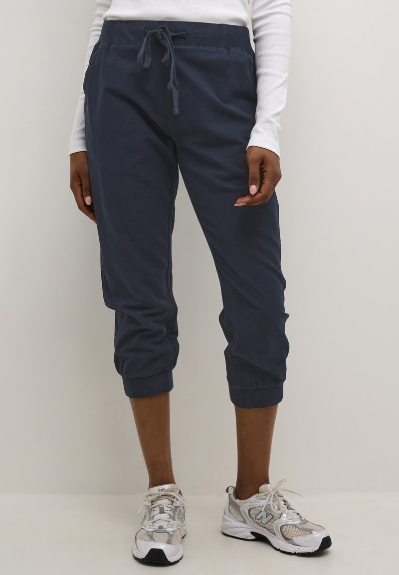 Femme portant un jogging court bleu marine, un haut à manches longues blanc et des baskets blanches, debout devant un fond uni.