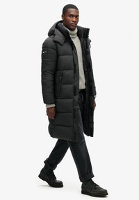 Abrigo largo negro tipo plumas con capucha, diseño acolchado, cierre delantero con cremallera y botones de presión, llevado sobre un suéter de punto gris claro y jeans oscuros.