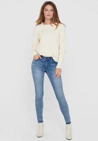 Cremefarbener Strickpullover mit langen Ärmeln, kombiniert mit hellblauen Skinny-Jeans und cremefarbenen Ankle-Boots. Einfaches Design und figurbetonte Silhouette.
