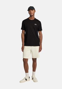Man i svart EA7 t-shirt, beige shorts, vita strumpor, svart och vit sneakers och en svart keps, stående mot en ljus, enhetlig bakgrund.