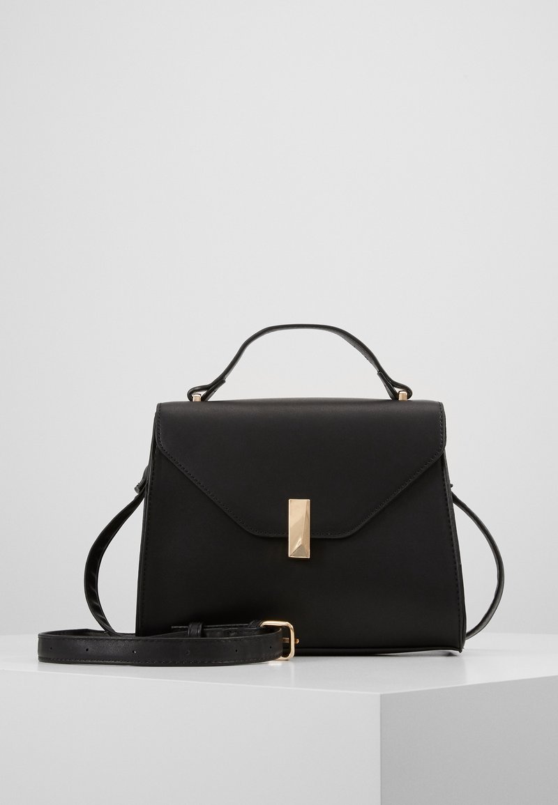 Sac à main en cuir noir avec poignée supérieure, bandoulière réglable et fermoir rectangulaire doré sur une surface blanche avec un fond gris clair.