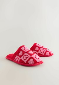 Červené chlpaté papuče s textúrovaným dizajnom obsahujúcim ružové srdcové vzory na kockovanom pozadí. Štýl s otvorenou špičkou a jemnou vnútornou výstelkou.