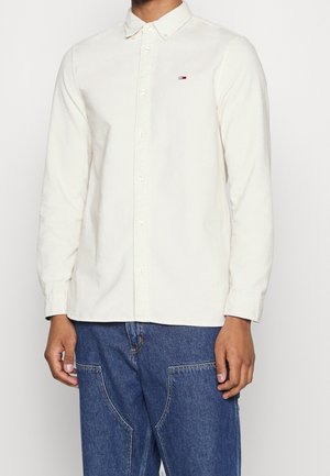 Mand iført en off-white skjorte med knapper og lange ærmer med et lille rødt, hvidt og blåt logo samt blå denim jeans.