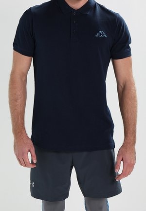 Mężczyzna w granatowej koszulce polo z logo Kappa, ciemnych sportowych szortach i szarych legginsach kompresyjnych stojący na jednolitym tle.