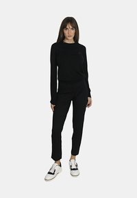 Maglione nero a maglia con maniche lunghe e un sottile logo rosso, abbinato a pantaloni neri slim e sneakers bianche con dettagli neri.