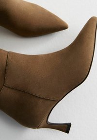 Stivali alla caviglia in suede marrone con punta appuntita e tacco sottile. La parte superiore presenta dettagli cuciti e una texture liscia.