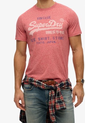 Mand iført rød SuperDry t-shirt og jeans med en ternet skjorte bundet om livet, læderbælte og armbåndsur, armene afslappede.