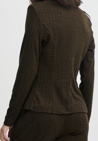 Femme portant un blazer marron à motif chevron et un pantalon assorti, vue de dos sur un fond uni.