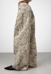 Osoba w szerokich spodniach w beżowo-szarym marmurkowym wzorze i czarnych, spiczastych butach, stoi na jednolitym tle.