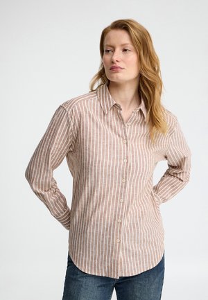 Femme aux cheveux roux portant une chemise boutonnée rayée verticalement marron clair et blanc et un jean bleu foncé, les mains dans le dos, regardant vers la droite.