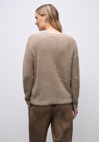 Beige lockerer Strickpullover aus weichem, strukturiertem Material mit weitem Ausschnitt. Kombiniert mit braunen, maßgeschneiderten Hosen, die eine entspannte Passform und Komfort zeigen.