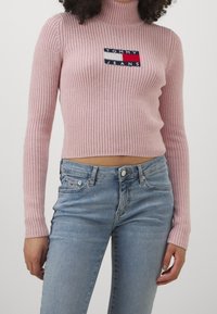 Rosa ribbad polotröja med en broderad logga i marinblått, rött och vitt. Kombinerad med ljus tvätt, högkända jeans.