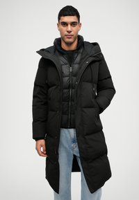 Čierny páperový kabát s overalovým a gombíkovým zapínaním, kapucňou a prešívanou textúrou. Nosený nad svetlejším páperovým kabátom, doplnený modrými džínsami.