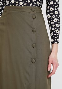 Olivgrüner Rock mit glatter Textur, ausgestattet mit fünf großen, runden Knöpfen vorne und einem hohen Taillendesign.