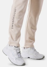 Ljust beige träningsbyxor med "SWEDEMOUNT"-logotyp på sidan, i kombination med vita Fila-sneakers med nät- och gummidetaljer.