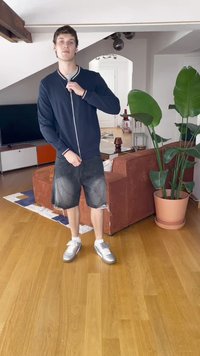 Tamsiai mėlyna zip-up striukė su baltu apykaklės kraštu, juodi džinsiniai šortai su nusidėvėjusiais kraštais ir sidabriniai sportbačiai. Vidinė aplinka su augalais ir baldais.