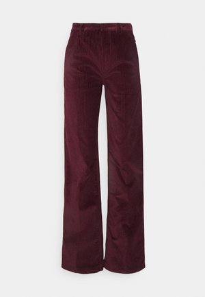 Nainen, jolla vihreä pitkähihainen paita avoimilla hihoilla, viininpunaiset corduroy-housut, beiget korkokengät ja beige clutch-laukku kädessä.