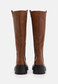 Paire de bottes hautes en cuir marron avec semelles noires épaisses, vues de dos, sur un fond blanc uni.