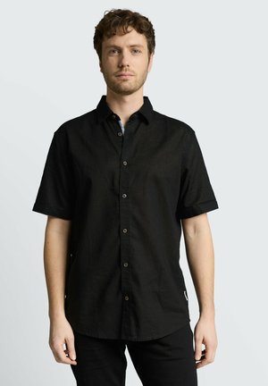 Junger Mann mit kurzen lockigen Haaren, der ein schwarzes kurzärmliges Hemd mit Knöpfen trägt, steht vor einem schlichten hellen Hintergrund.