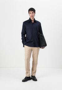 Cămașă bleumarin cu nasturi, pantaloni bej adaptați, pantofi negri, ținând o geantă din piele neagră. Material neted, croială bine definită și design minimalist.
