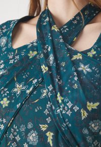 Il tessuto floreale turchese presenta un mix di fiori bianchi, gialli e blu. Il design include una scollatura aperta e dettagli drappeggiati.