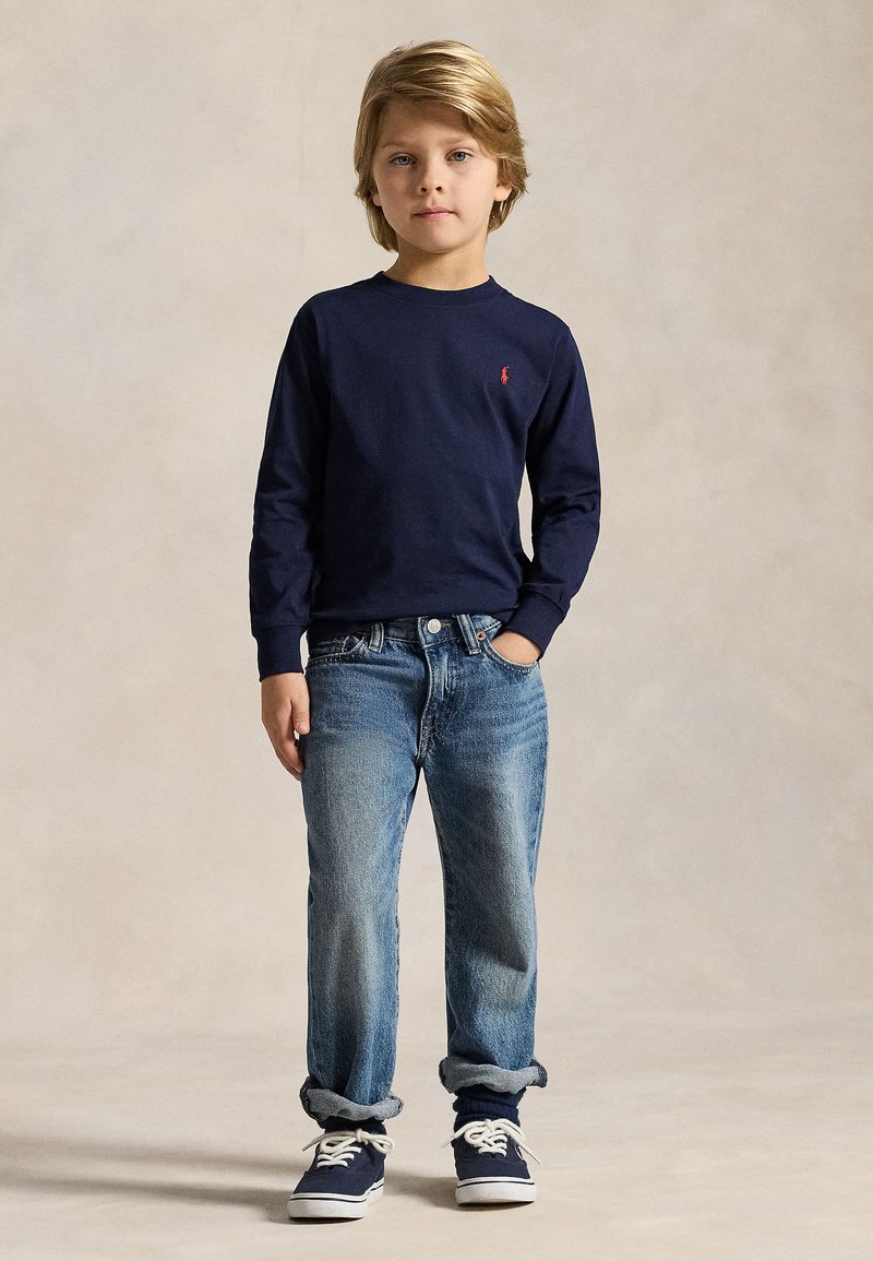 En ung dreng med blondt hår iført en marineblå langærmet skjorte, blå jeans med opsmøgede ben og marineblå sneakers, der står mod en ensfarvet baggrund.
