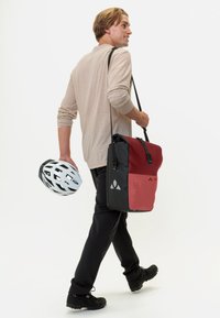 Borsa a tracolla nera e rossa con tracolla regolabile, forma rettangolare, materiale resistente, accenti grafici e design elegante. Uomo che tiene un casco bianco.