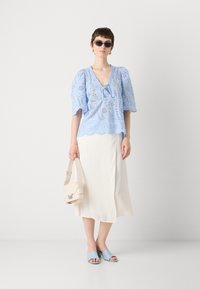 Lichtblauw kanten blouse met pofmouwen, V-hals en gegolfde zoom. Gecombineerd met een witte midi-rok, lichtblauwe instappers en een beige tas.
