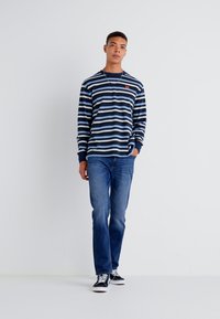 Long-sleeve navy and white striped shirt, lightweight fabric, round neckline, small logo on chest, paired with blue jeans and black sneakers.
