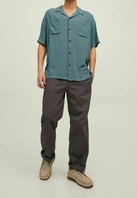 Uomo che indossa una camicia button-up a maniche corte color turchese, pantaloni grigio scuro e scarpe beige con lacci, in piedi con una mano in tasca.