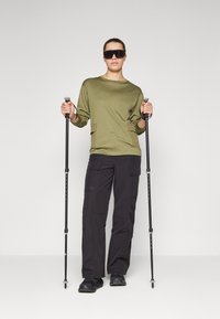 Olive green long-sleeve top with two front pockets, paired with black cargo pants. Black hiking poles in each hand, black footwear.