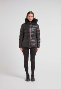 Czarna kurtka puchowa z kapturem wykończonym futrem, z zamkiem z przodu, ribbowanymi akcentami i dwiema bocznymi kieszeniami. Noszona na czarnych legginsach i botkach.