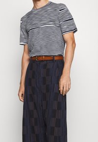 Hombre vestido con una camiseta a rayas en azul marino y blanco, con pantalones anchos de cuadros en azul oscuro y un cinturón marrón, de pie con una mano a su lado.