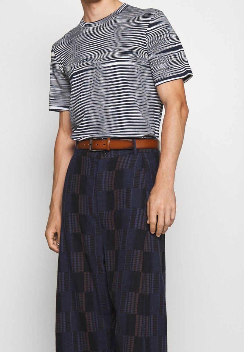 Hombre vestido con una camiseta a rayas en azul marino y blanco, con pantalones anchos de cuadros en azul oscuro y un cinturón marrón, de pie con una mano a su lado.
