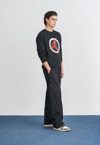 Μαύρο πουκάμισο με μακρυά μανίκια και κόκκινο γραφικό λογότυπο, συνδυασμένο με μαύρα παντελόνια με κόκκινες ραφές. Το μοντέλο φοράει γκρι και λευκά παπούτσια.