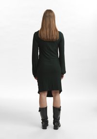 Žalia ilga rankovė su aukšta-žema apatine dalimi, pagaminta iš lygaus audinio. Suderinta su juodais kelio ilgio botais su sagtimis ir tekstūruota paviršiumi.