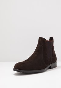 Hnědé suede kotníkové boty s elastickými bočními panely, kulatou špičkou a nízkým vrstveným podpatkem, s jemným šitím a texturovaným akcentem.