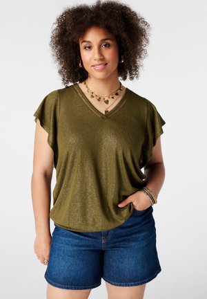 Femme aux cheveux bouclés portant un haut vert olive à manches courtes, des colliers dorés, des bracelets et un short en jean bleu, une main dans la poche.