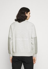 Felpa grigia chiara con finitura testurizzata, dotata di maniche lunghe, polsini a coste e dettaglio testuale bianco sulla parte posteriore.