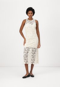 Kanten jurk in off-white met bloemenpatronen, zonder mouwen en een golvende zoom, aangevuld met zwarte platte schoenen.