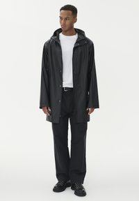 Fekete vízálló esőkabát kapucnival, patent gombokkal és hosszú ujjakkal, fekete nadrággal és fekete cipővel párosítva, semleges háttér előtt.