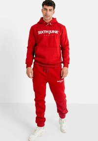 Rode fleecetrui met capuchon en bijpassende joggingsbroek met witte logo. Beschikt over een voorzak, een trekkoordcapuchon en geribbelde boorden. Sneakers maken de look compleet.