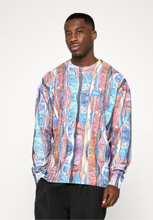 Mann iført fargerik, mønstret langermet skjorte med vertikale striper og svarte bukser, står foran en ensfarget hvit bakgrunn.