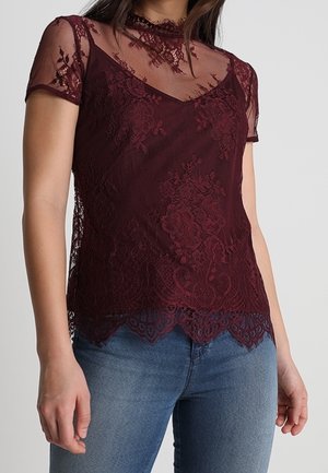 Bordowa koronkowa bluzka z krótkimi, przejrzystymi rękawami, ozdobiona kwiatowymi wzorami i falowanym dołem. Warstwowy fason z dopasowanym krojem.