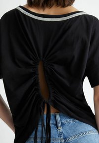 T-shirt nera con un design arricciato sul retro, caratterizzata da un'apertura ovale e un colletto adornato da strass. Tessuto morbido e maniche corte.