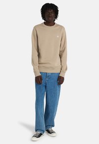 Young man standing wearing beige crewneck sweatshirt, loose blue jeans, and black-and-white sneakers on plain light background.