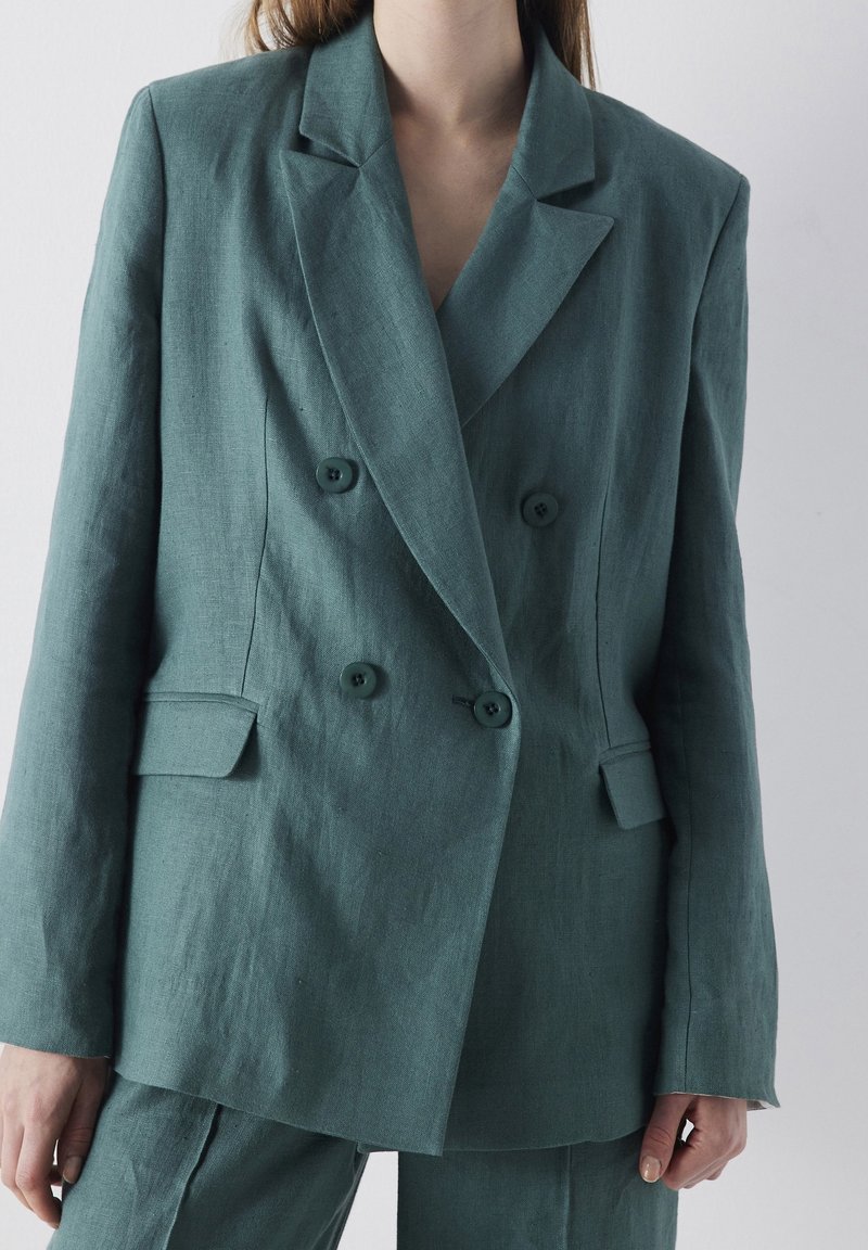 Femme portant un blazer en lin bleu sarcelle croisé et un pantalon assorti sur un fond clair uni.